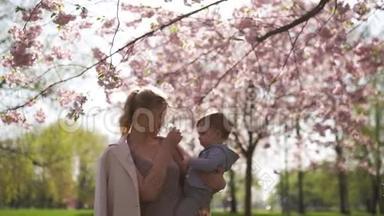 年轻的妈妈抱着她的小儿子，在樱花盛开的樱花树下，飘落的粉红色花瓣和